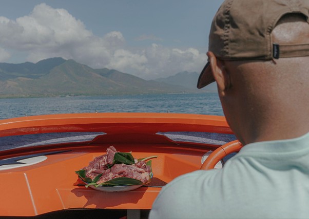 adha pengorbanan 15 dompet dhuafa Back of a person wearing a cap on an orange boat, plate of sliced meat with greens on the console, ocean and mountains in the distance.