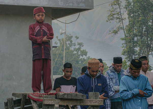adha pengorbanan 2 dompet dhuafa Young boy in maroon traditional clothing stands on a wooden platform, while a group of men watch beside a gray building outdoors against a mountainous backdrop.