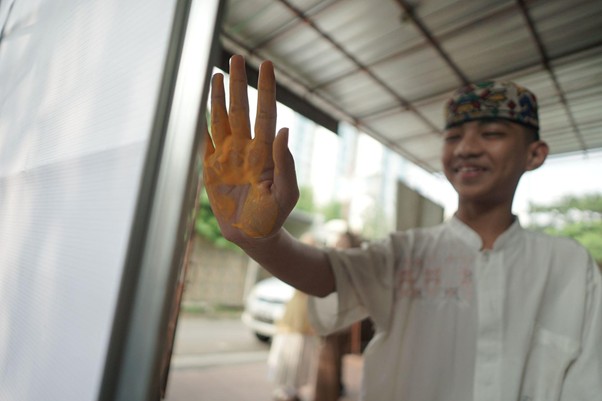 Young man in a traditional cap and white shirt presses an orange-painted hand against a glass window, smiling.