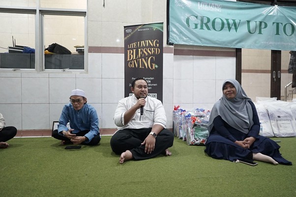 Man in a white shirt speaks into a microphone while seated on a green carpet, flanked by a boy in blue and a woman in a hijab at a charity event.