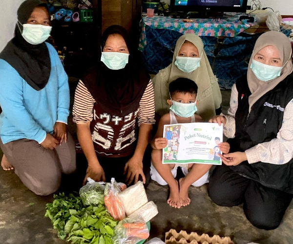 fotojet 1 dompet dhuafa Five masked people, including a child, sit together indoors while the child holds a nutrition certificate; fresh vegetables and eggs are laid out in front.