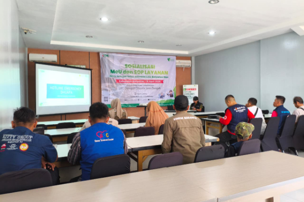 Training room with participants seated facing a projector screen; a banner at the front reads 'Sosialisasi Menu dan SOP Layanan' with a presenter at the table.