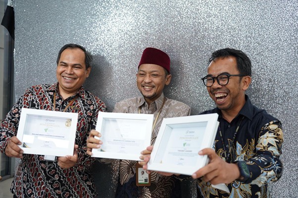 penghargaan penghimpunan filantropi indonesia 1 dompet dhuafa Three men in batik shirts smile while holding framed certificates, standing against a silver speckled wall; center man wears a red cap (peci).