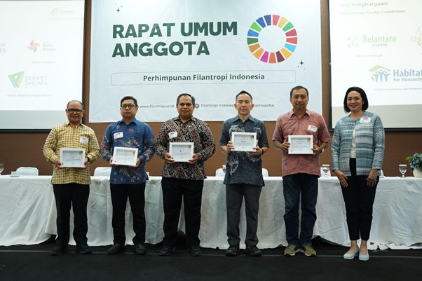 penghargaan penghimpunan filantropi indonesia 10 dompet dhuafa Group of six men and one woman on stage holding certificates at an awards ceremony, with a banner that says 'RAPAT UMUM ANGGOTA' in the background.