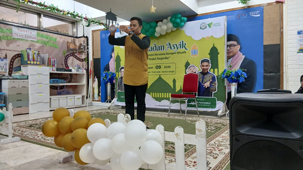 ramadan asyik kb ra istiqlal 2 dompet dhuafa Man in a mustard shirt speaks into a microphone on a decorated stage with white and gold balloons and a Ramadan-themed banner in the background (indoors).