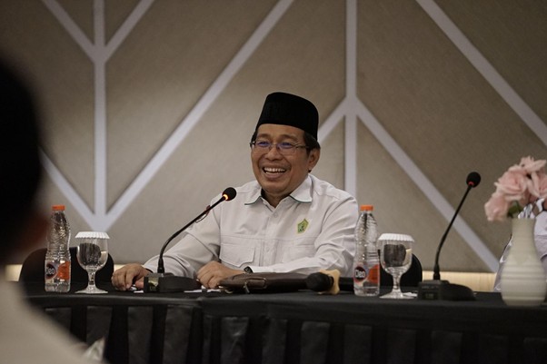 sk laz 2026 5 dompet dhuafa Smiling man in a white shirt and black cap sits at a panel table with microphones, water bottles, and flowers at a conference.
