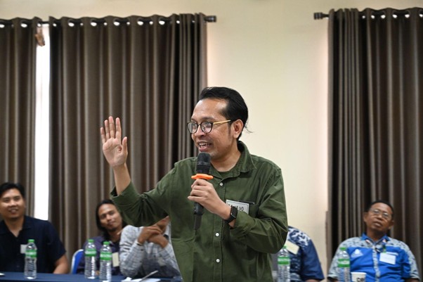training of trainer ik dd 4 dompet dhuafa Man in green shirt speaks into a microphone while raising his left hand in a conference room, addressing a panel of people seated at a table with water bottles.