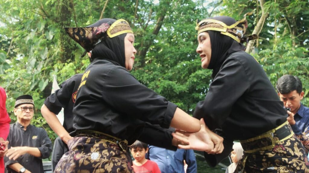 whatsapp image 20260427 at 141122 dompet dhuafa Two women in black traditional outfits and ornate batik sashes perform a Indonesian dance, holding hands across a green park setting while onlookers watch.