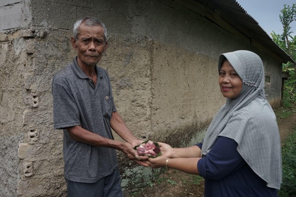yuyum peternak berdaya 8 dompet dhuafa An elderly man and a woman wearing a headscarf exchange a small bag of vegetables outside a rural mud-walled house.