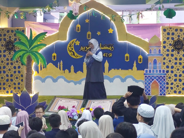 zakat fitrah sekolah embun pagi 2 dompet dhuafa A woman in a hijab speaks into a microphone on a colorful stage, addressing a crowd of students.