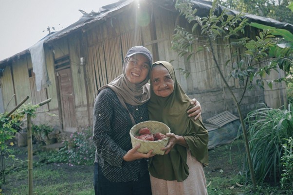 Two women in headscarves smile and embrace while holding a basket of red produce in a garden in front of a wooden house.