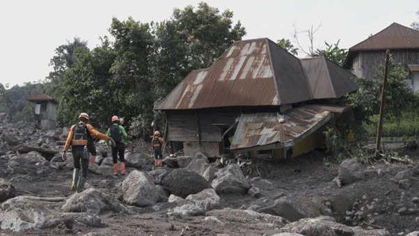 Rescue workers traverse a rocky debris field beside a collapsed, rusted-roof house after a natural disaster.