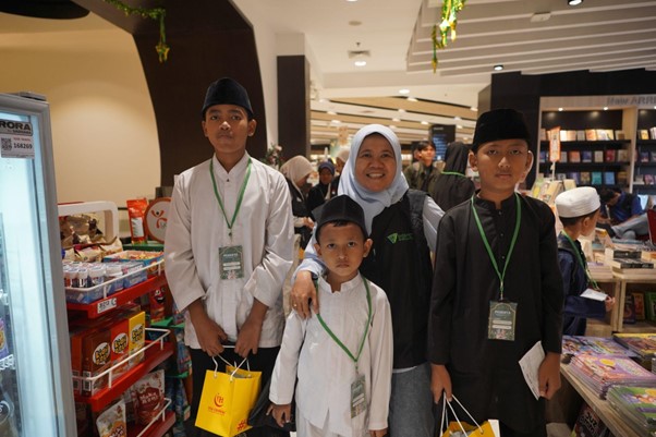 50anakyatimbelanjagramediabsd04 dompet dhuafa Family of five in traditional white clothing with green lanyards posing in a bookstore or library, holding gift bags.