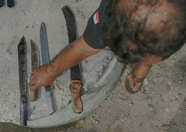 adhapengorbanan11 dompet dhuafa Top-down view of a man in flip-flops inspecting several knives and blades laid out on a curved surface on a dusty ground.