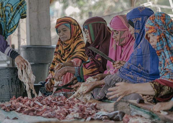 adhapengorbanan14 dompet dhuafa Women in colorful headscarves kneeling or seated around a table, cutting and sorting raw meat with knives at an outdoor market or communal prep area.