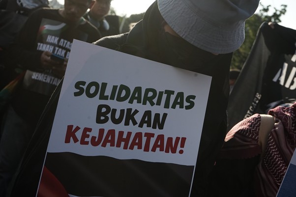 aksi kamisan pembebasan relawan gsf 4 dompet dhuafa Protester wearing a hat and face covering holds a sign that reads 'SOLIDARITAS BUKAN KEJAHATAN!'