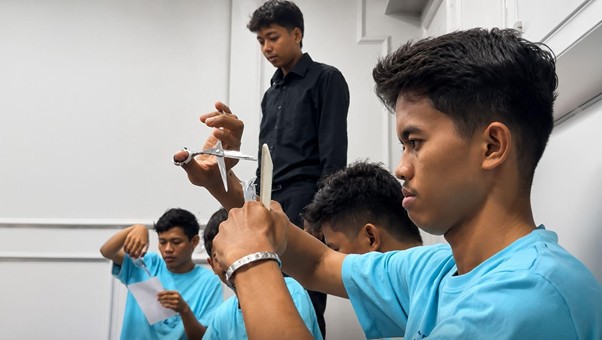 Young men in a bright classroom workshop practice knife skills, supervised by an instructor; foreground student handles a training knife with focus.