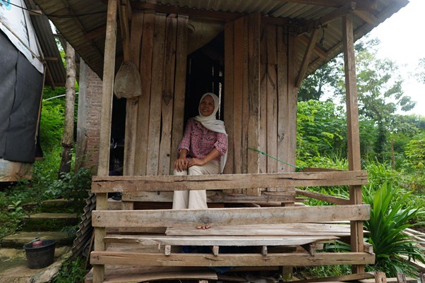 bangunasadarikandangkambing2 dompet dhuafa Woman wearing a hijab sits on the wooden front porch of a rustic hut, with a green garden in the background.