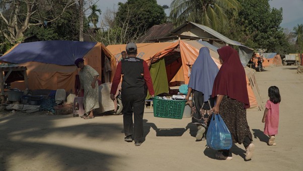 bantuankamiideaaceh2 dompet dhuafa People walking along a row of orange tents in a sandy camp; a man in dark clothing leads, with women in colorful robes and a child nearby carrying bags.