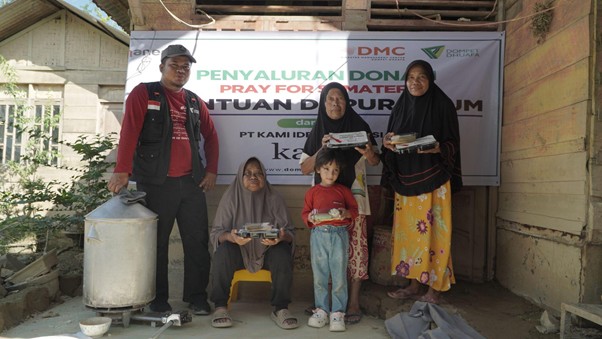 bantuankamiideaaceh3 dompet dhuafa Family of five and a volunteer pose with food packages at a relief donation site, banner behind them, and a large water container on the left.