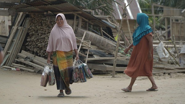 bantuankamiideaaceh4 dompet dhuafa Two women walk along a dirt path carrying multiple plastic shopping bags, with wooden structures in the background.