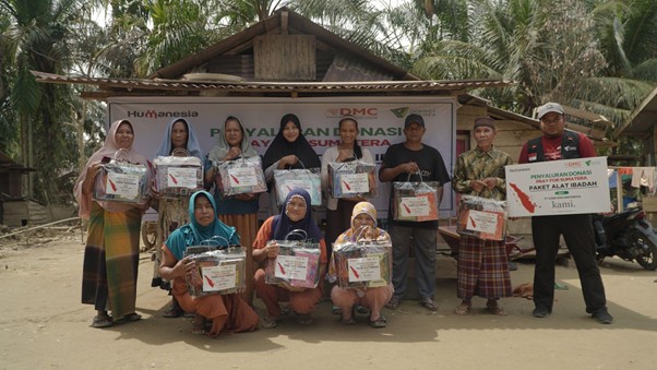 bantuankamiideaaceh5 dompet dhuafa Group of volunteers posing outdoors with relief packages in clear plastic bags; some kneel in front, a banner and palm trees visible in the background.