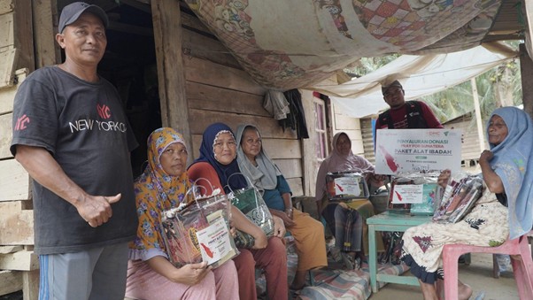 bantuankamiideaaceh7 dompet dhuafa Man stands beside a row of seated villagers receiving gift packages outdoors under a striped canopy.