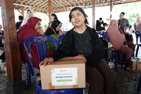 bantuanmodenauntukibuibutunanetra7 dompet dhuafa Woman seated under a wooden pavilion holds a donation box labeled 'Penyaluran Donasi Berbagi Sembako' as others sit on blue chairs in the background at a charity gathering.