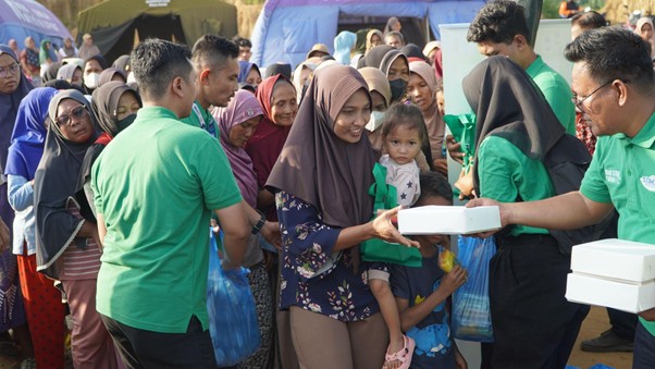 bantuanstimunountukacehtamiang5 dompet dhuafa People line up to receive food boxes from volunteers in green shirts at an outdoor aid event; a mother in a hijab holds her child as she accepts a box.