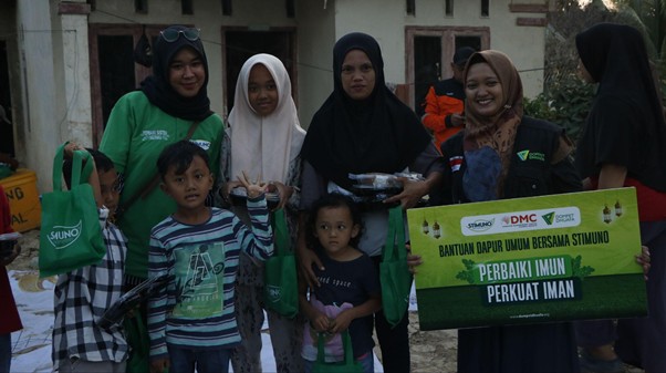 bantuanstimunountukacehtamiang6 dompet dhuafa Group of Indonesian women and children pose with green tote bags at a community aid event, a banner in the background promoting immunization and togetherness.