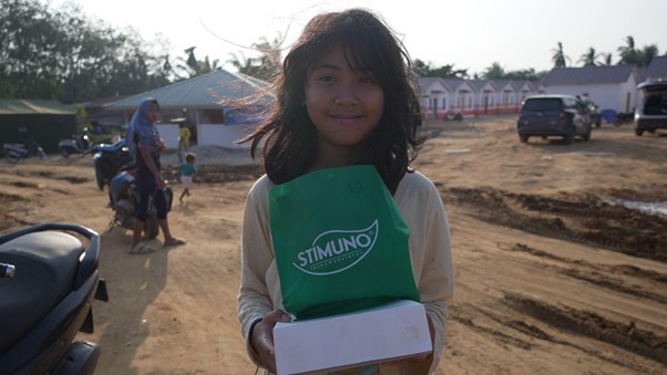 bantuanstimunountukacehtamiang8 dompet dhuafa Girl in a rural area holding a white box, wearing a green Stimuno vest and a smile; houses and dirt road in the background.