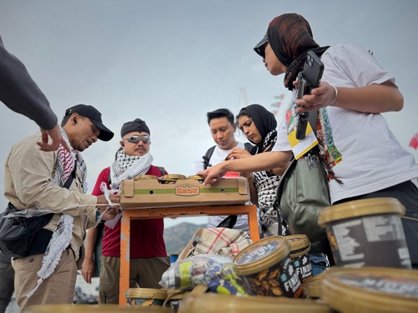 bantuan logistik untuk palestina 1 dompet dhuafa Group of people gathered around a wooden crate on a table, inspecting items at an outdoor event, with cans and bags in the foreground.