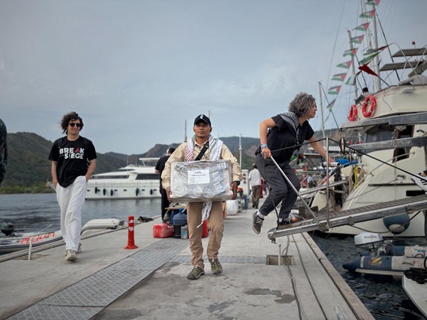 bantuan logistik untuk palestina 2 dompet dhuafa People on a marina dock with luxury yachts; a man in a tan suit carries a large wrapped box, while two others walk and climb toward a boat.