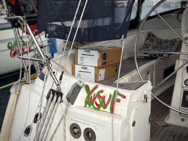 bantuan logistik untuk palestina 3 dompet dhuafa Boxes labeled humanitarian aid stacked on a sailboat cockpit beside the steering wheel, with rigging lines and winches in the foreground.
