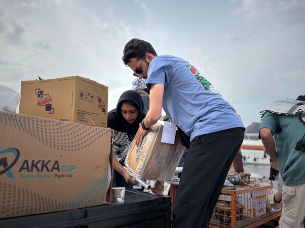 bantuan logistik untuk palestina 4 dompet dhuafa Volunteer in a blue T-shirt helps a woman in a hijab unload a large box from a truck at a relief drive outdoors.