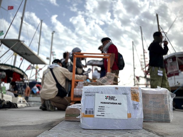 bantuan logistik untuk palestina 5 dompet dhuafa Box labeled humanitarian aid on a dock with people sorting supplies and boats in the background