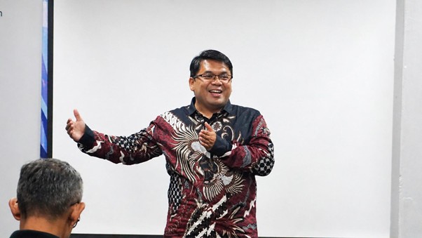 Smiling man in a batik shirt giving a presentation with arms open to the audience in a classroom or meeting room.