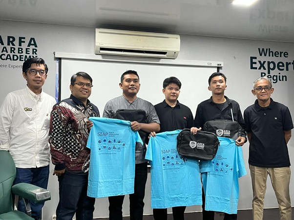 Six men stand in a training room holding light-blue T-shirts with logos, a whiteboard and projector screen behind them.
