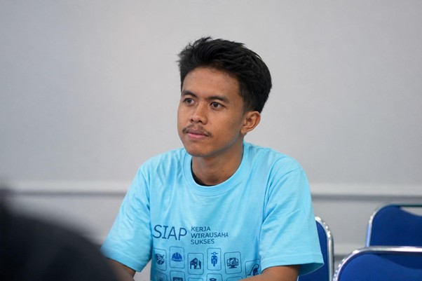 Young man with short black hair and a mustache sits at a table, wearing a light blue SIAP T‑shirt, in a room with blue chairs.