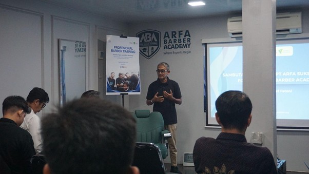 Instructor speaks to an audience in a barber training room at ARFA Barber Academy, with a poster and projector displaying slides.
