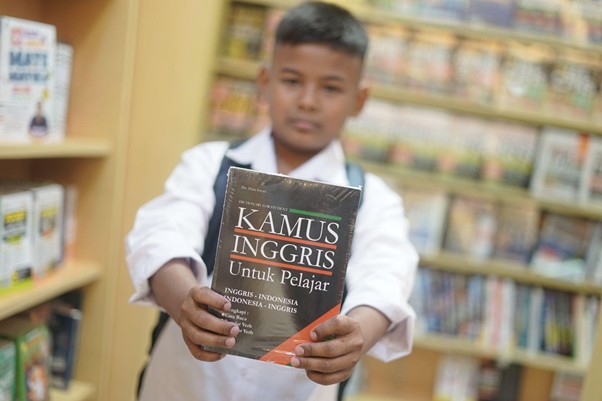 belanjabarengyatimgramediadanptrajarasakuliner16 dompet dhuafa Boy in a white shirt with a backpack holds up a bilingual English–Indonesian dictionary titled 'Kamus Inggris Untuk Pelajar' toward the camera in a library.