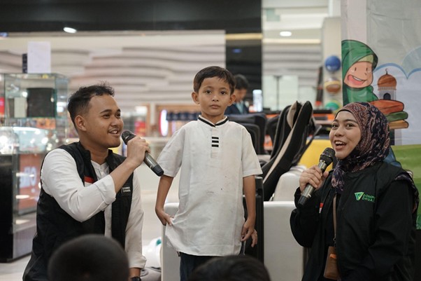 belanjabarengyatimgramediadanptrajarasakuliner17 dompet dhuafa Man on the left and a woman on the right hold handheld microphones while a boy in a white shirt stands between them during an indoor event at a mall.