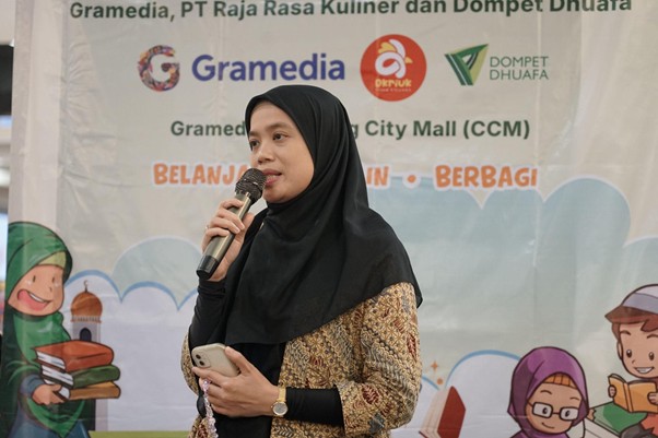 belanjabarengyatimgramediadanptrajarasakuliner18 dompet dhuafa Woman in a black hijab speaks into a microphone while holding a phone, standing before a banner with logos and cartoon students behind her.