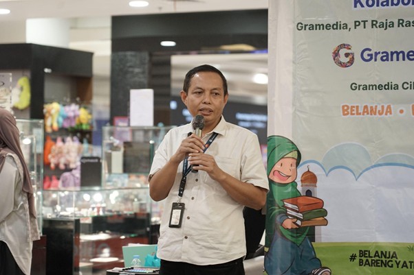belanjabarengyatimgramediadanptrajarasakuliner19 dompet dhuafa Man in a white short-sleeve shirt speaks into a microphone at a promotional booth, with a banner and shop displays in the background.