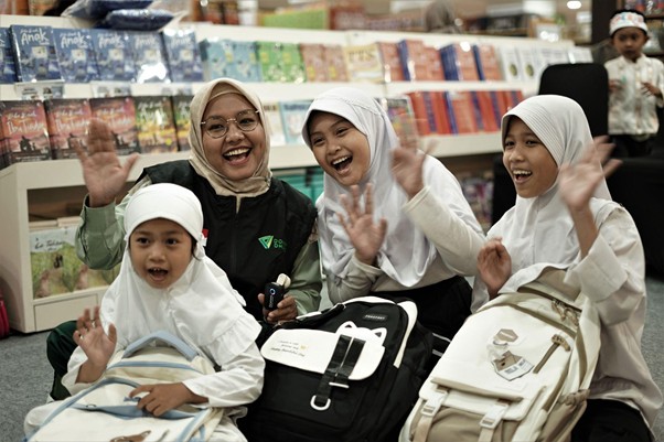 belanjabarengyatimnadia4 dompet dhuafa A joyful group of four girls in hijabs waving and smiling while sitting with backpacks in a bookstore or library aisle.