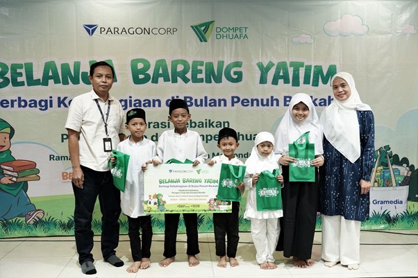 belanjabarengyatimparagon2 dompet dhuafa Group of children in white outfits and two adults pose on a stage, each holding green bags, at a charity event with the banner 'Belanja Bareng Yatim' in the background.