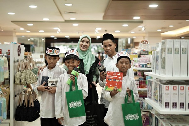belanjabarengyatimparagon3 dompet dhuafa Mother and father with four children posing in a bright store, the kids wearing white shirts, 'Experi' hats, and holding bags and products.
