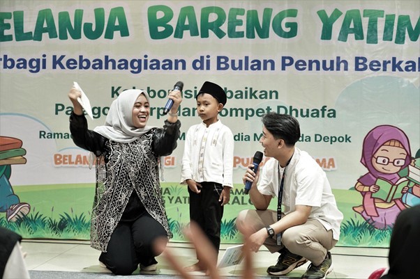 belanjabarengyatimparagon5 dompet dhuafa Woman in hijab laughing, a boy in a cap, and a man with a microphone on stage at a charity event with a colorful banner.