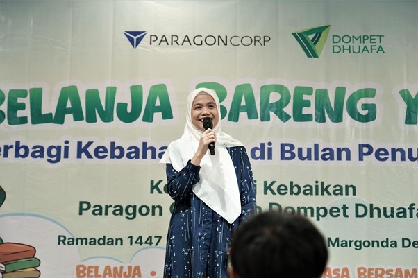 belanjabarengyatimparagon7 dompet dhuafa Woman in a white hijab and navy dress speaks into a microphone on stage at a charity event banner backdrop showing ParagonCorp and Dompet Dhuafa logos.