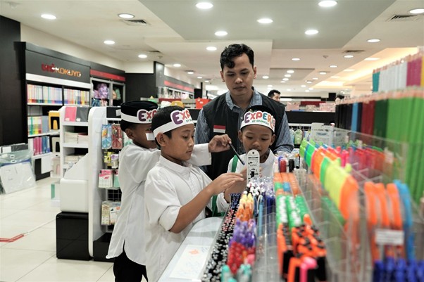 belanjabarengyatimparagon9 dompet dhuafa Four children in a stationery store, guided by an adult, as they examine products at a colorful marker display on the aisle.